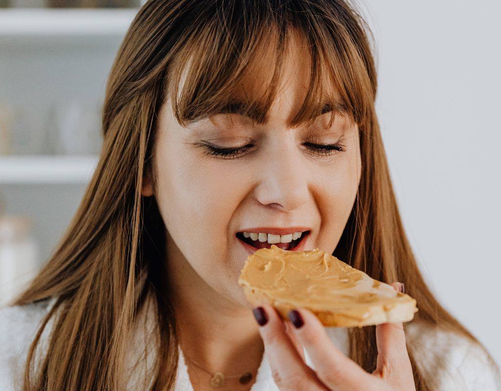 Mulher mordendo pão com pasta de amendoim e apreciando os benefícios da pasta de amendoim para saúde