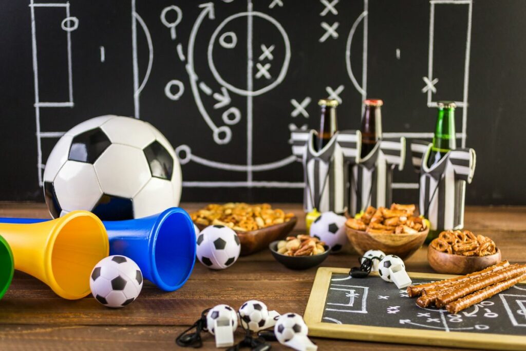 Imagem com comidas, bolas de futebol e vuvuzela em cima da mesa, e um esquema tático de fundo.