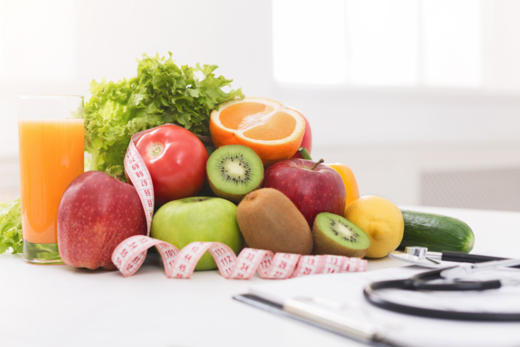 Mesa de um nutricionista com frutas, salada, suco, estetoscópio, prancheta e fita métrica.