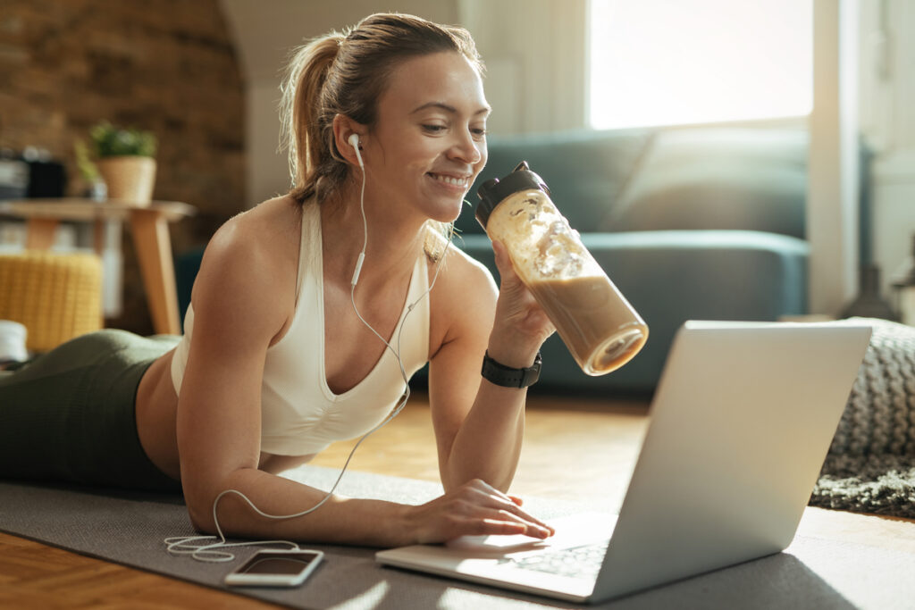 Jovem desportista feliz bebendo whey protein enquanto relaxa no chão e navega na net no laptop